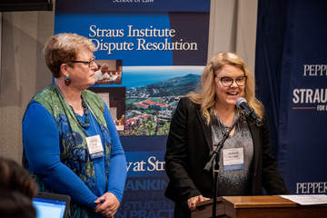 two woman stand at a podium while one speaks at a Straus event