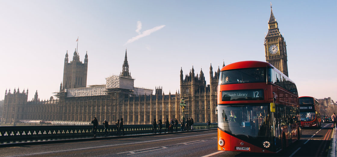 a red bus in London crossing a bridge with parliament in the background