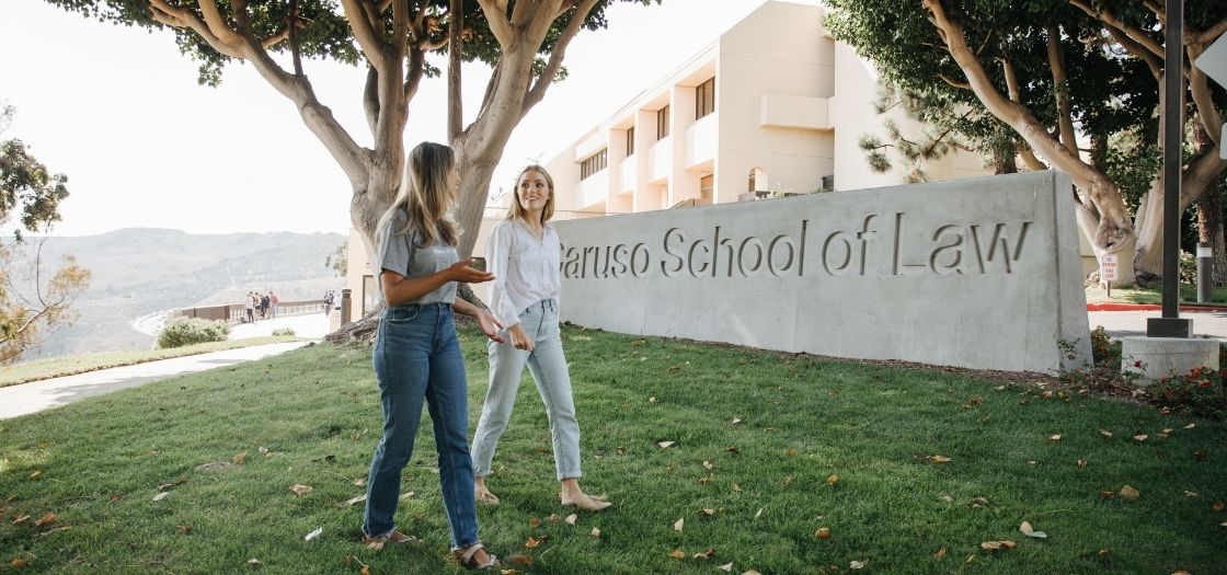 Two female students walking in the foreground of the Caruso School of Law sign