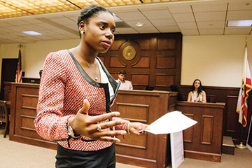 A female student stands in the Trial Courtroom holding a paper and presenting a case 