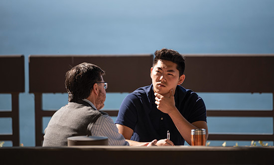 an asian male student sits on the terrace speaking with a male professor