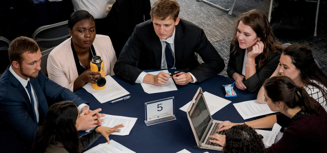 Seven students work on laptops at a table wearing suits