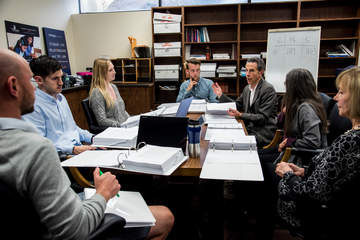a group of seven people sit around a table full of papers having an animated discussion