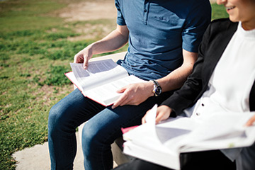 Two students sitting on a bench outside flipping  through textbooks