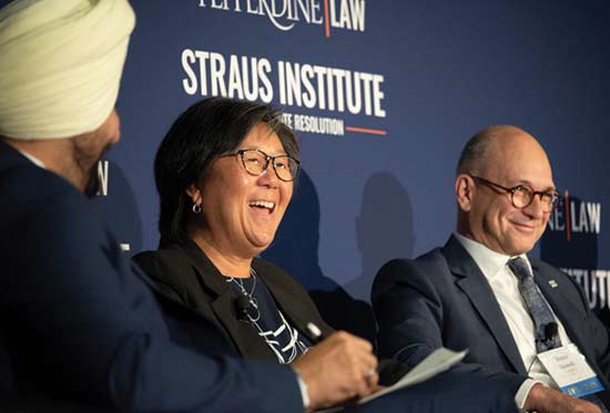 A woman laughing with men seated on either side of her in front of a Straus Institute blue sign