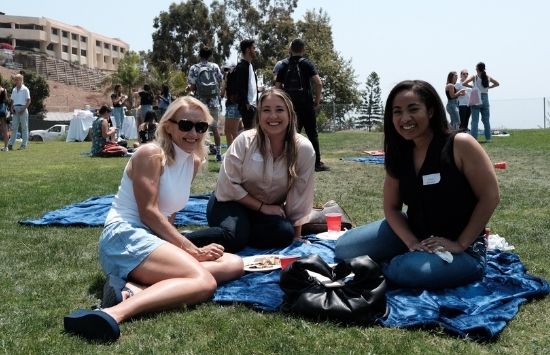 Three female students sit on a blue blanket at the BBQ